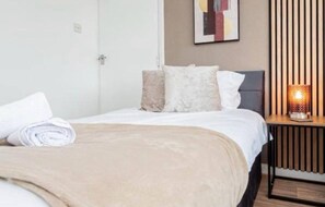 A neatly made bed with a beige blanket and pillows, a bedside table with a lamp, and a wall with a framed abstract painting. - Stylish 3BR Home Near Nec, Bhx, HS2 & JLR (Birmingham)