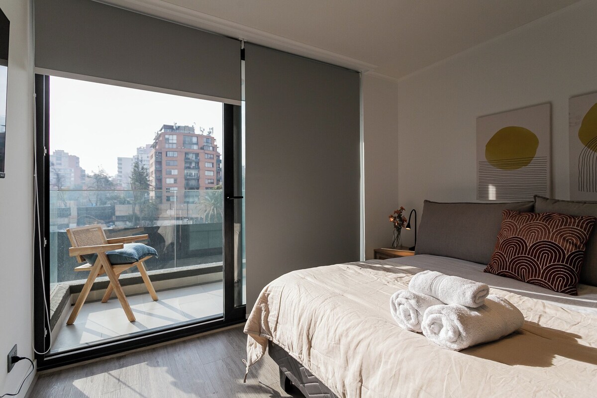 A modern bedroom with a large bed, a balcony with a chair, and a city view.