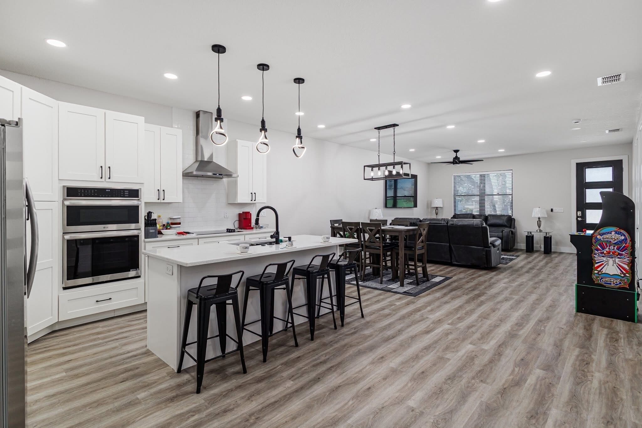 A modern kitchen with white cabinets, a central island with bar stools, and a dining area with a black sofa and chairs.