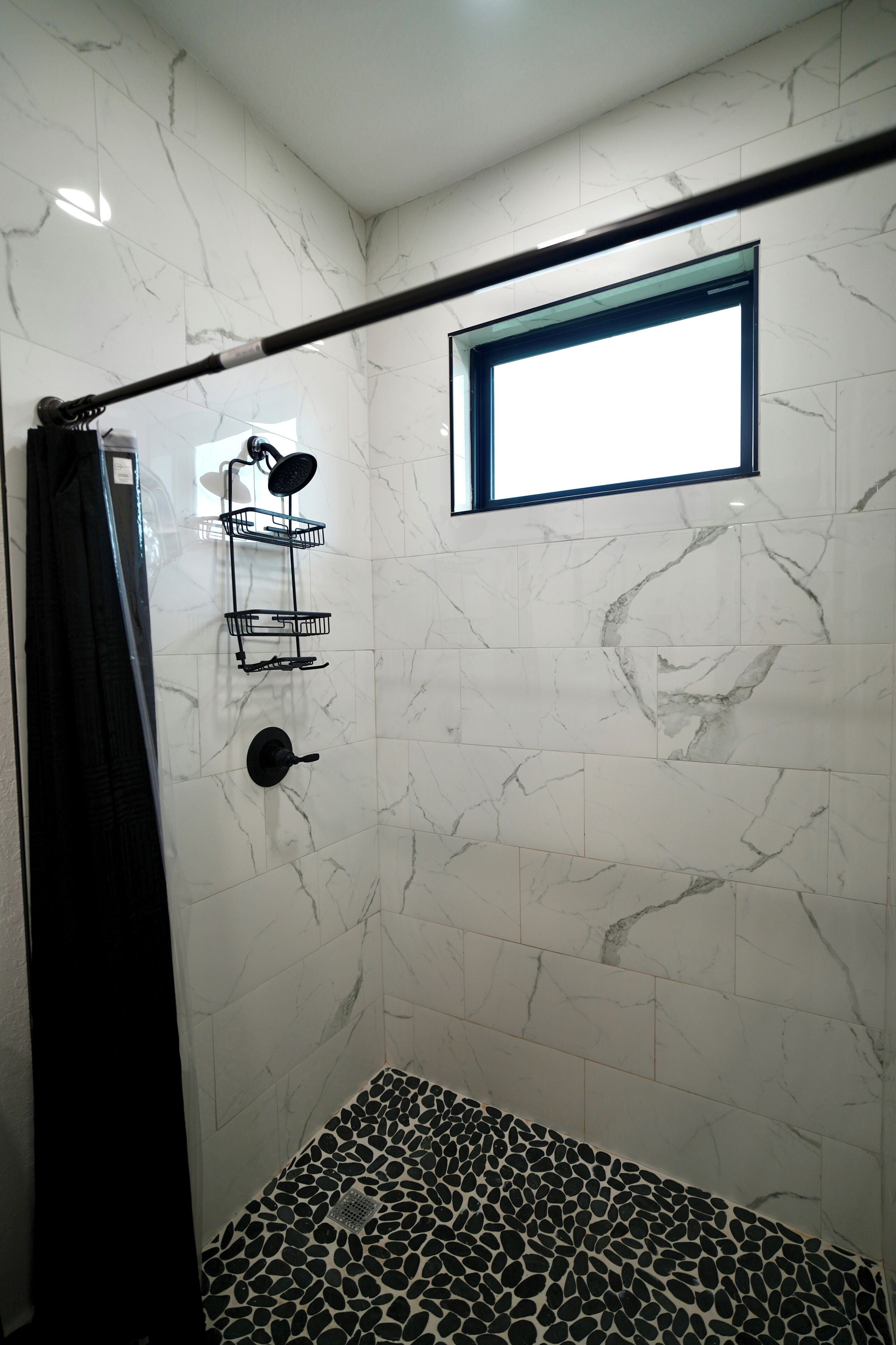 A shower area with a black shower curtain, a small window, and a tiled floor with a pebble pattern.