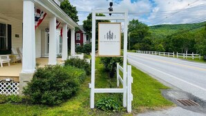 A white building with a porch, a signboard, and a road with a white fence in the background. - Bridgewater Living - Woodstock Suite (Bridgewater)