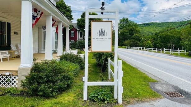 A white building with a porch, a signboard, and a road with a white fence in the background.