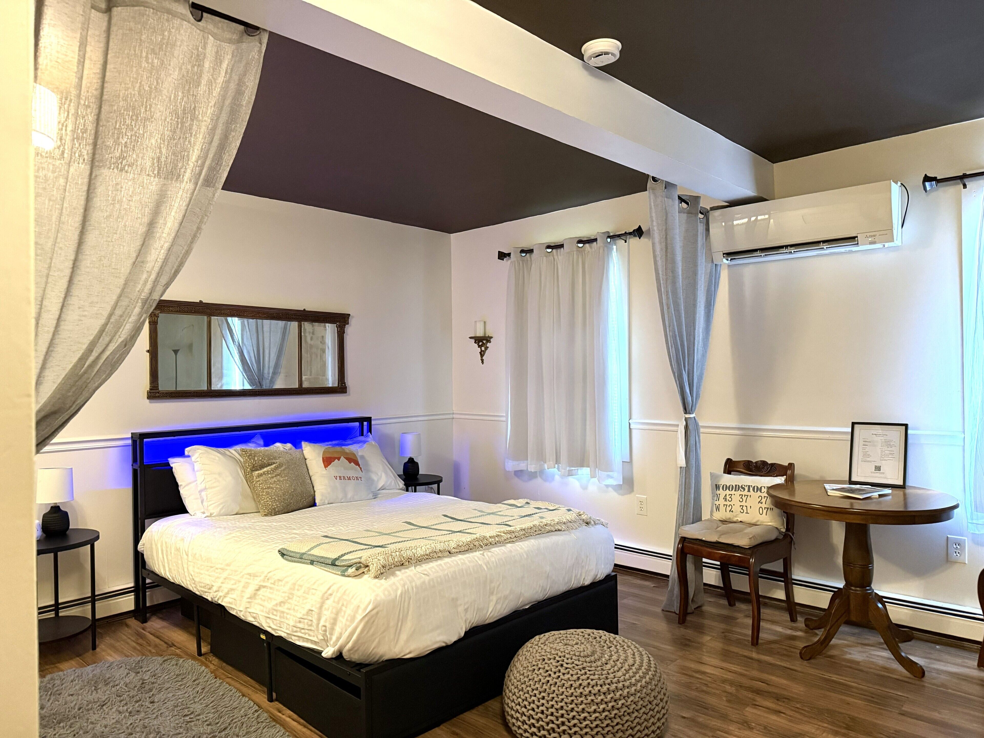 A bedroom with a bed, bedside tables, a round wooden table, a chair, a mirror, and an air conditioning unit.