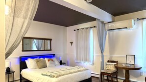 A bedroom with a bed, bedside tables, a round wooden table, a chair, a mirror, and an air conditioning unit.