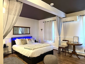 A bedroom with a bed, bedside tables, a round wooden table, a chair, a mirror, and an air conditioning unit.
