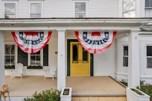 A two-story house with a porch decorated with American flags. There are white chairs on the porch and a yellow door. - Bridgewater Living - Woodstock Suite (Bridgewater)