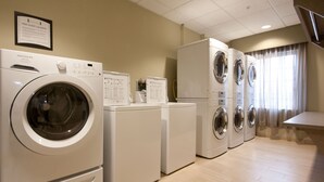 Une buanderie équipée de plusieurs machines à laver et de plusieurs sèche-linge, avec une pancarte indiquant « buanderie à disposition gratuite des clients » et un sol carrelé.