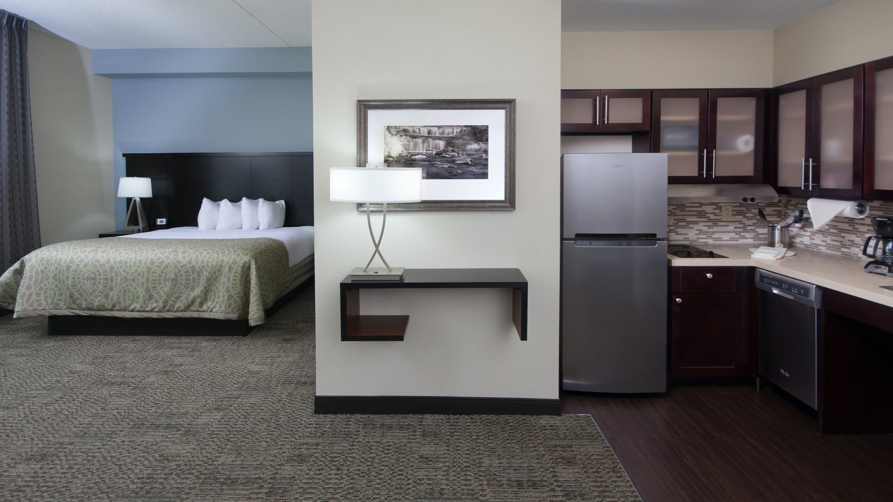 A hotel room with a bed, bedside table, lamp, picture frame, and a kitchenette area with a refrigerator and coffee maker.