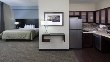 A hotel room with a bed, bedside table, lamp, picture frame, and a kitchenette area with a refrigerator and coffee maker.