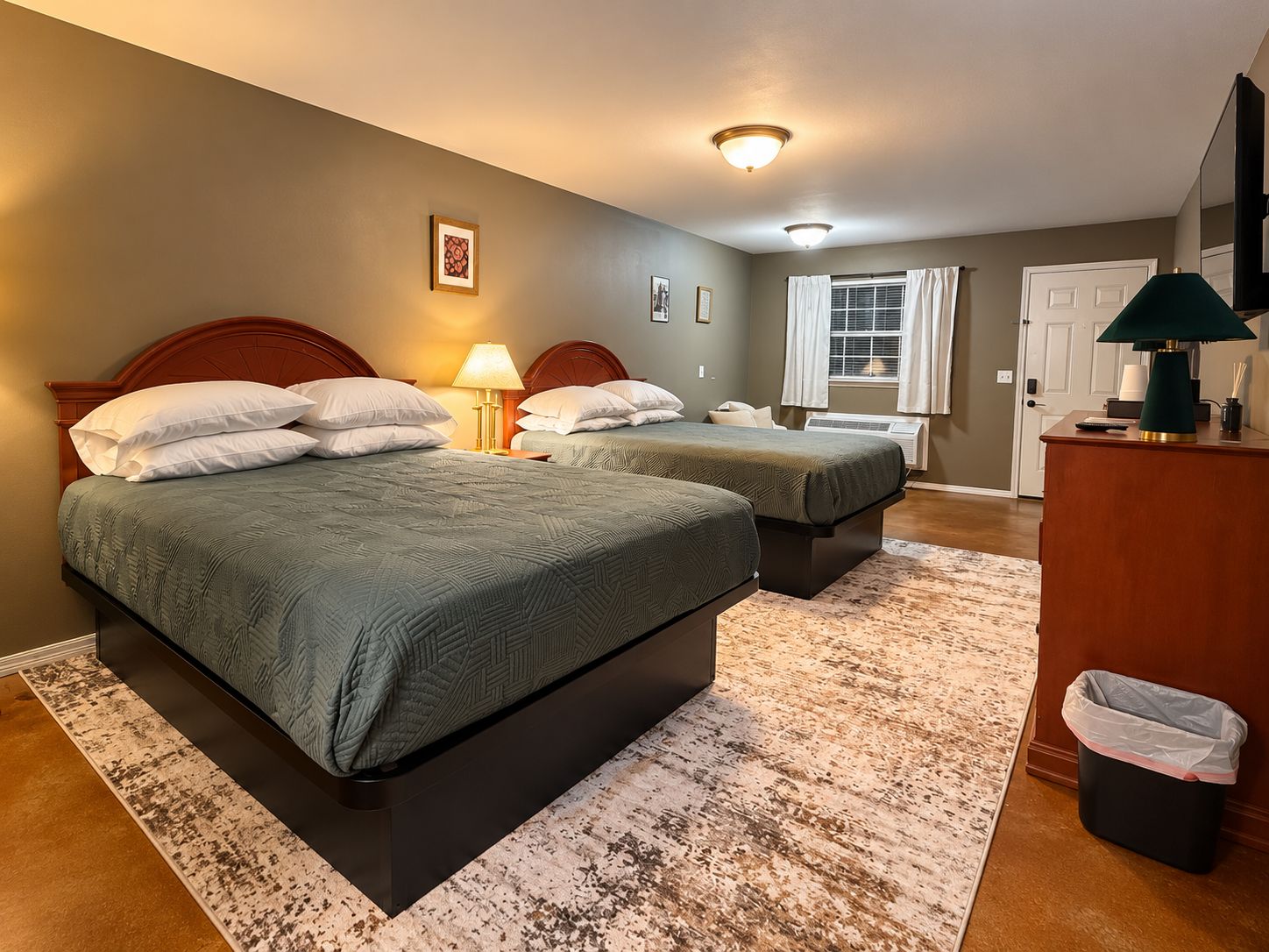A hotel room with two beds, a desk, a trash bin, and a window with curtains.
