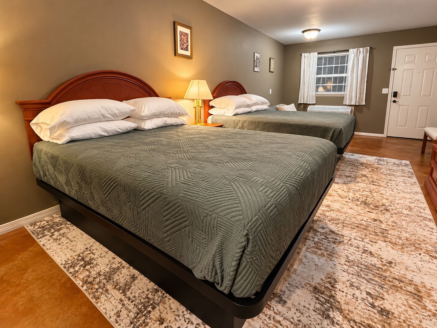 A bedroom with two beds, a nightstand, a lamp, a window with curtains, and a door.