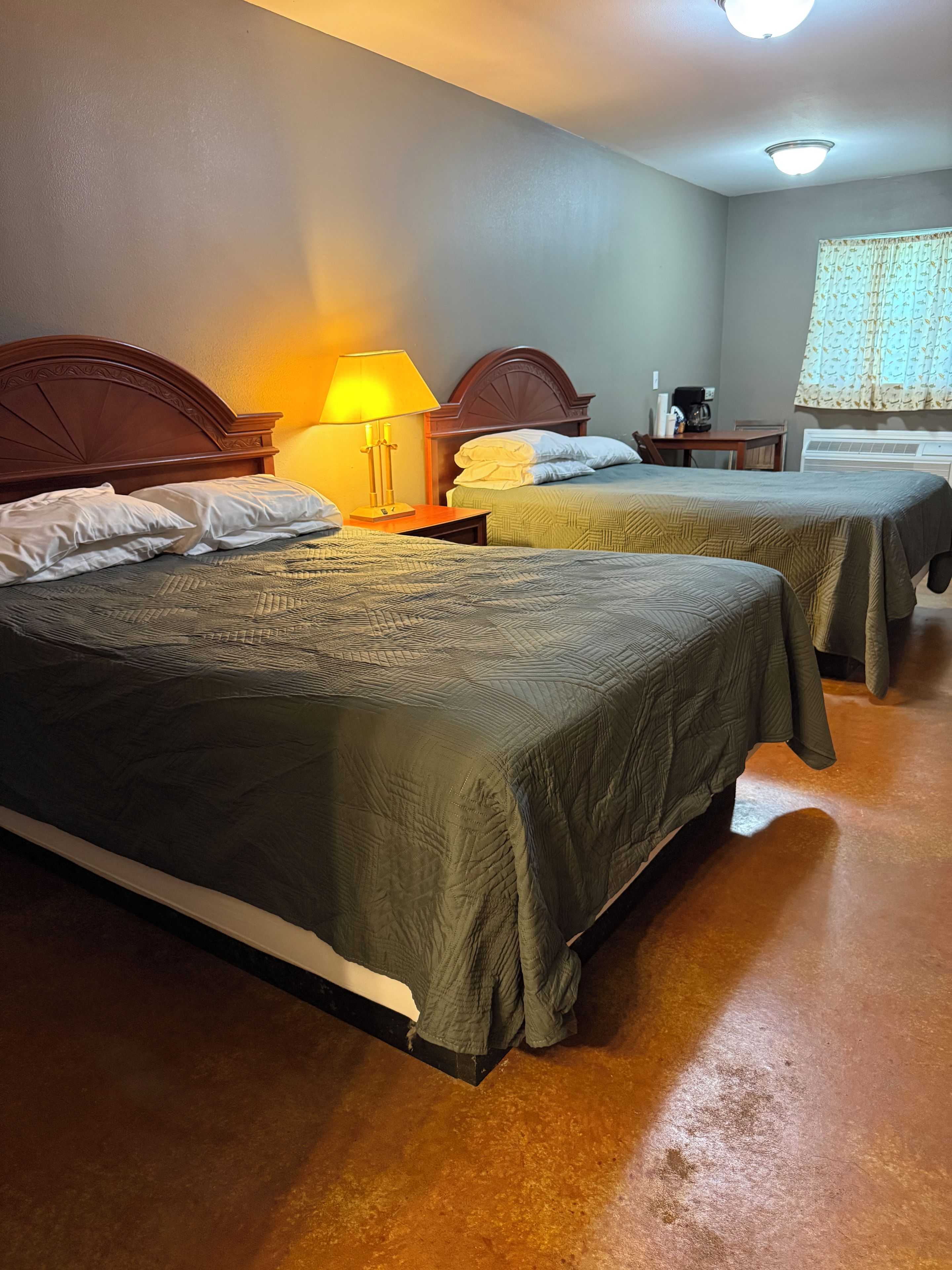 A hotel room with two beds, a nightstand with a lamp, a small table with a coffee maker, and a window with curtains.