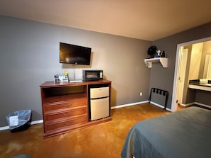 A compact hotel room with a bed, a desk with a microwave, a small refrigerator, and a television on the wall. - The Grove 3 — Rustic River Lodge Room (TAHLEQUAH)