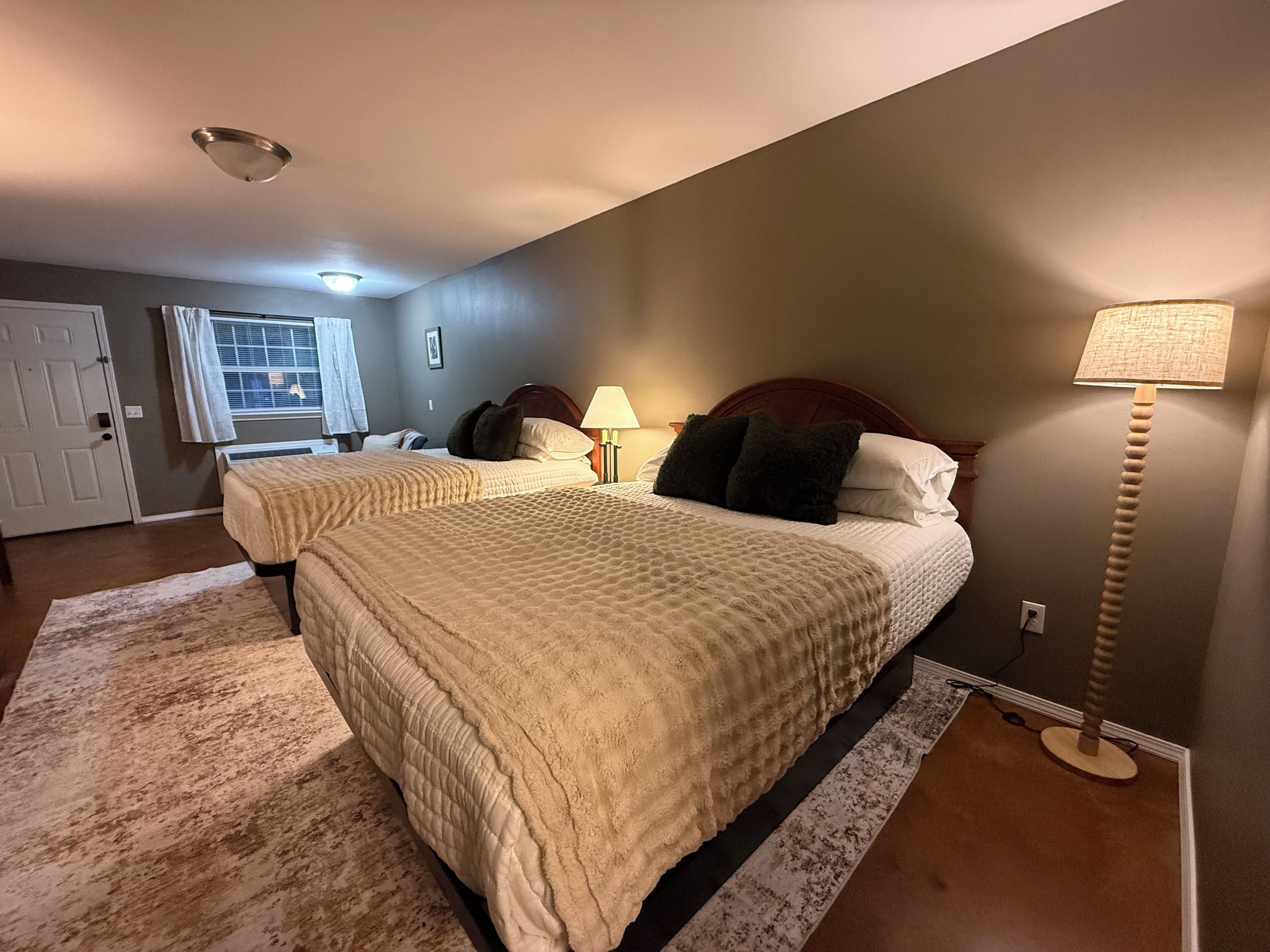 A bedroom with two beds, a window with curtains, a door, and a lamp.