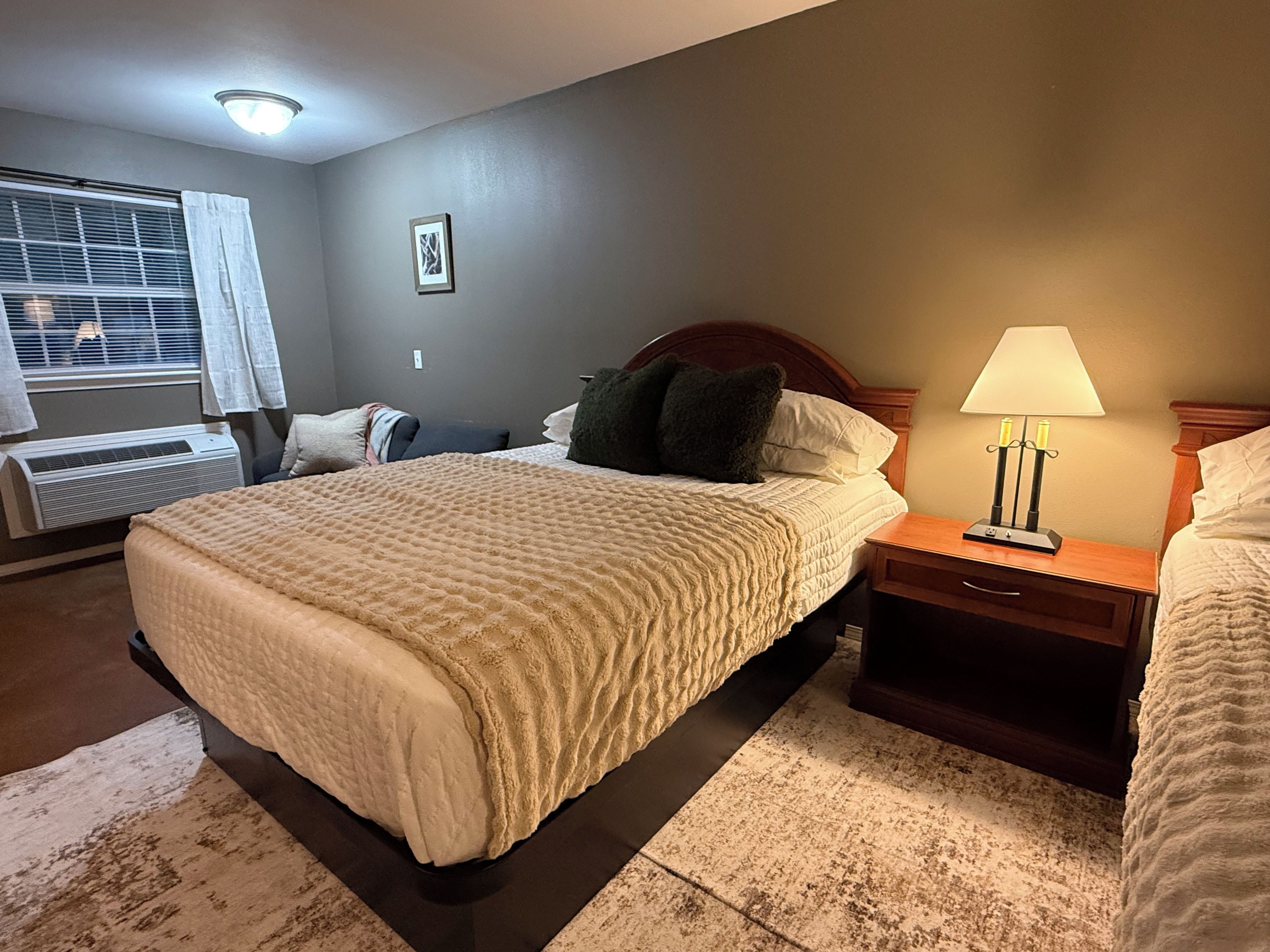 Habitación de hotel con una cama grande, mesitas de noche, una lámpara, una ventana con persianas y un equipo de aire acondicionado.