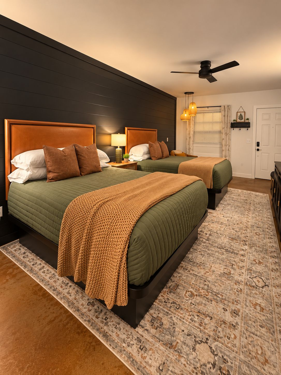 A hotel room with two beds, a ceiling fan, a bedside lamp, a patterned rug, and a door.