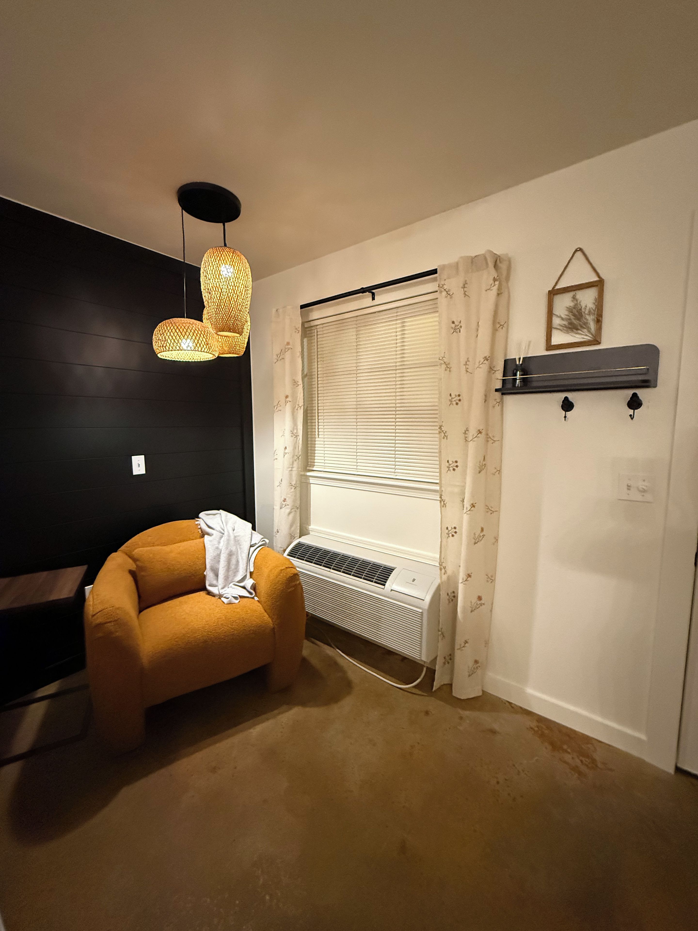 A room with a dark wall, a beige carpet, a small orange chair, a window with blinds, a hanging light fixture, and a wall-mounted shelf with a picture frame.