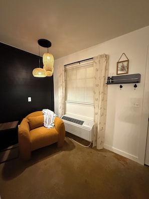 A room with a dark wall, a beige carpet, a small orange chair, a window with blinds, a hanging light fixture, and a wall-mounted shelf with a picture frame. - The Pines 1 — Updated River Lodge (TAHLEQUAH)