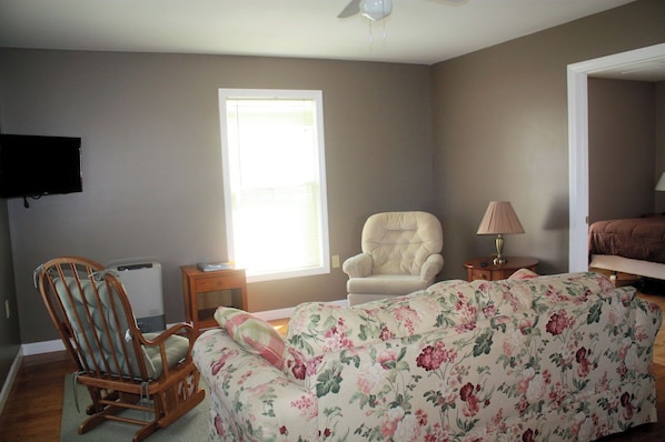 A room with a rocking chair, a floral sofa, a television, and a window with blinds.