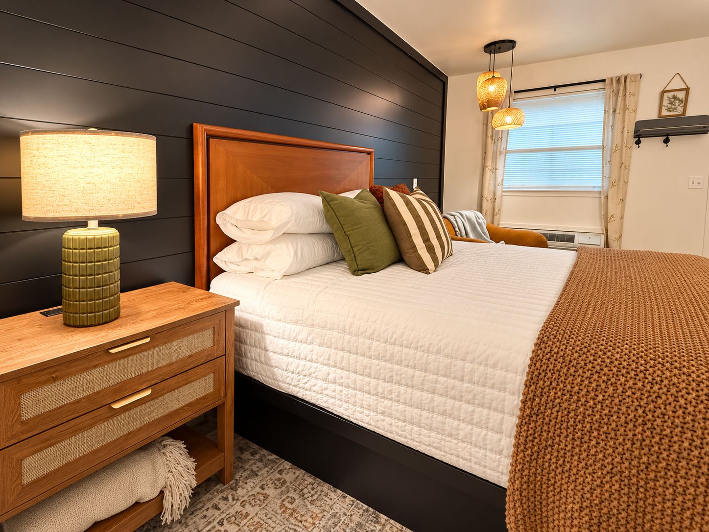 Un dormitorio con cabecera de madera, una mesita de noche con lámpara, cama con ropa blanca y varios cojines, una ventana con persianas y un aire acondicionado instalado en la pared.