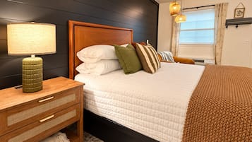Un dormitorio con cabecera de madera, una mesita de noche con lámpara, cama con ropa blanca y varios cojines, una ventana con persianas y un aire acondicionado instalado en la pared.