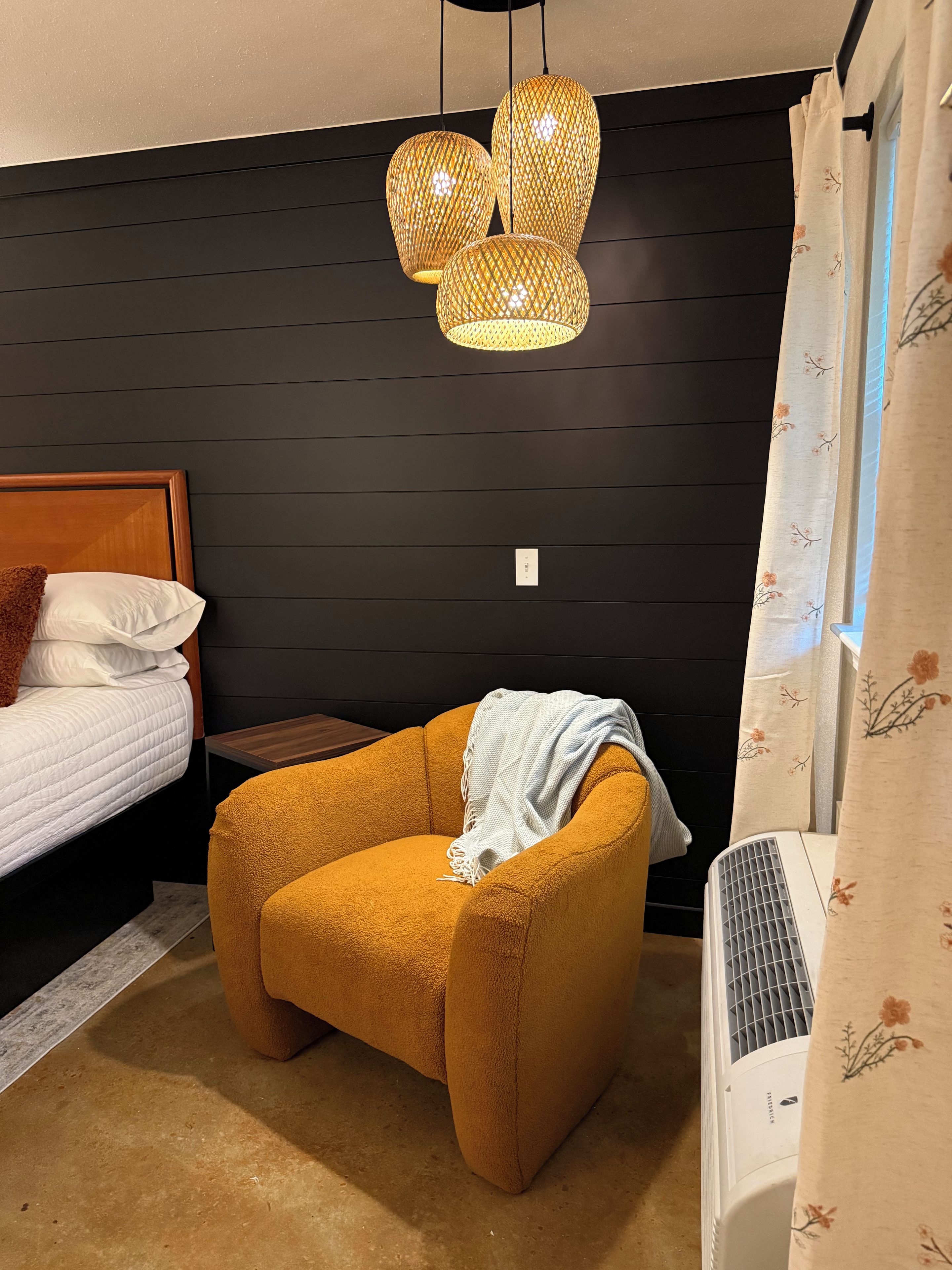 Un dormitorio con cama, una mesita, una silla, un calefactor y una lámpara fijada a la pared.