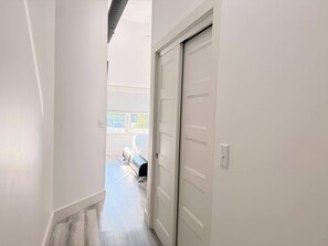 Um corredor com uma porta fechada, uma janela com persianas e uma cama ao fundo.