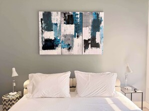 Quarto de hotel com uma cama, duas almofadas brancas, uma mesa de cabeceira com abajur e um quadro abstrato na parede.