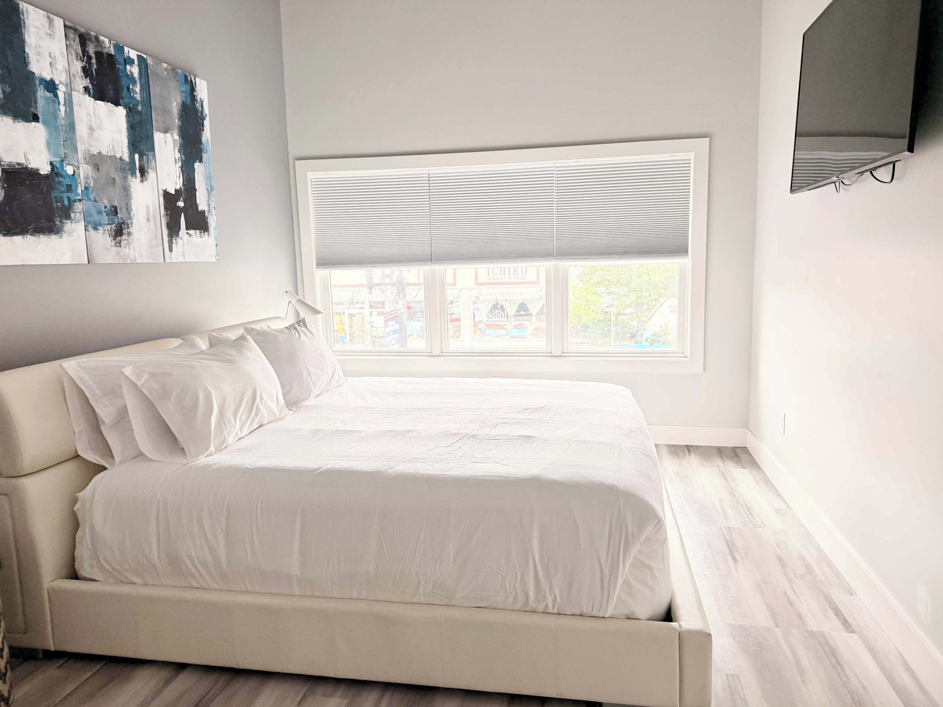 A bedroom with a bed, a window with blinds, a wall-mounted TV, and abstract art.