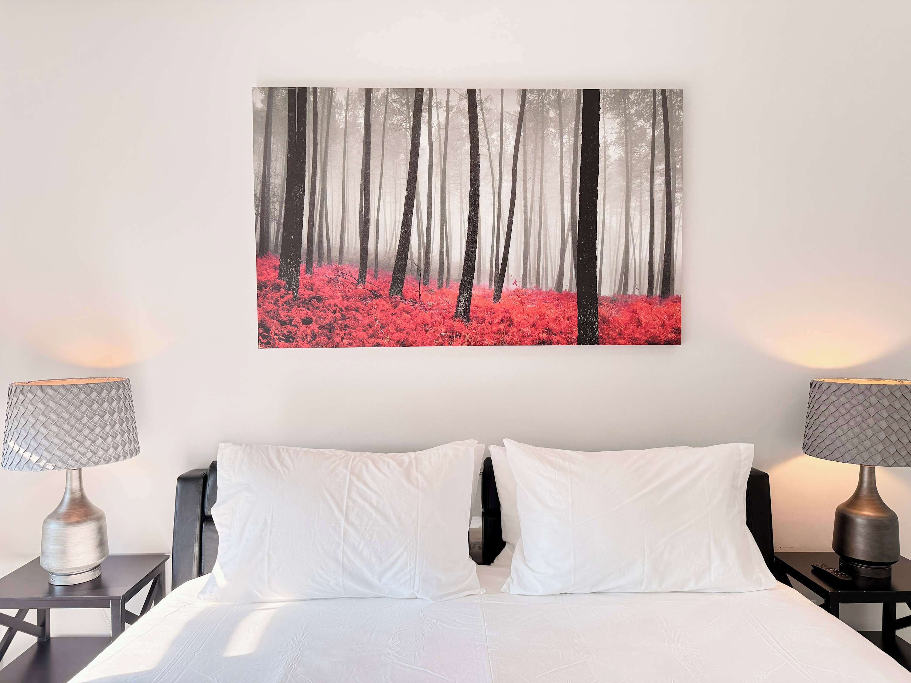 A bedroom with a bed, two bedside tables, and a wall-mounted painting of a forest.