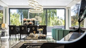 A modern living room with a piano, a dining area with chairs and a table, and a large window with a view of greenery.