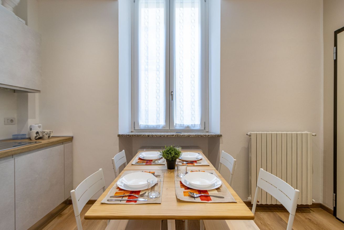 Ein Essbereich mit einem Holztisch, gedeckt für vier Personen, weißen Stühlen, einem Topfpflanzen und einem Fenster mit Vorhängen.