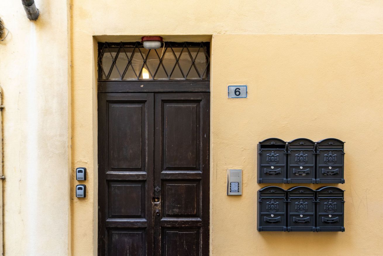 Una porta in legno scuro con una griglia metallica, una cassetta delle lettere con il numero 6 e due cassette postali fissate al muro.