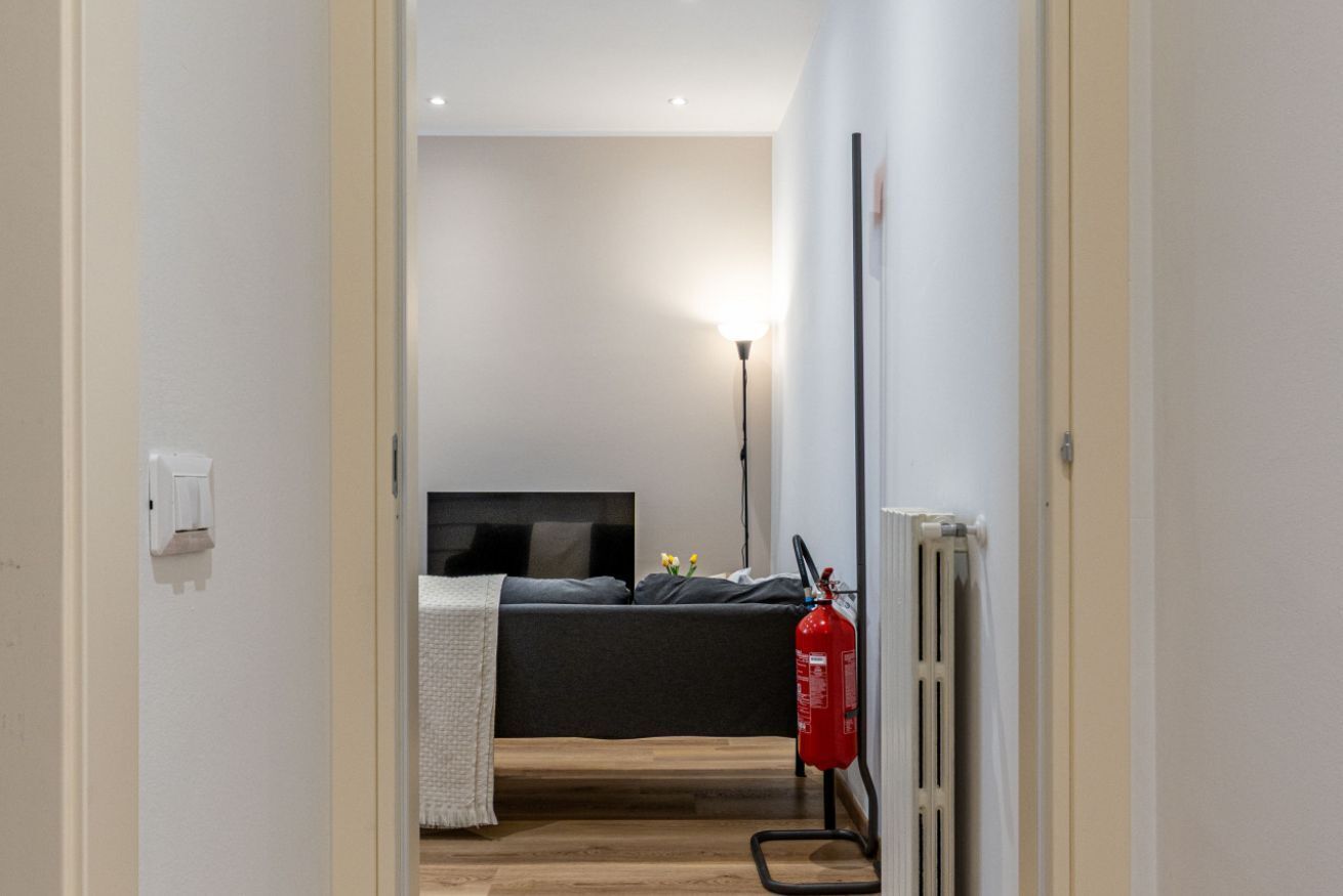 Ein schmaler Flur führt zu einem Schlafzimmer mit einem Bett, einem Nachttisch, einer Lampe und einem Feuerlöscher.