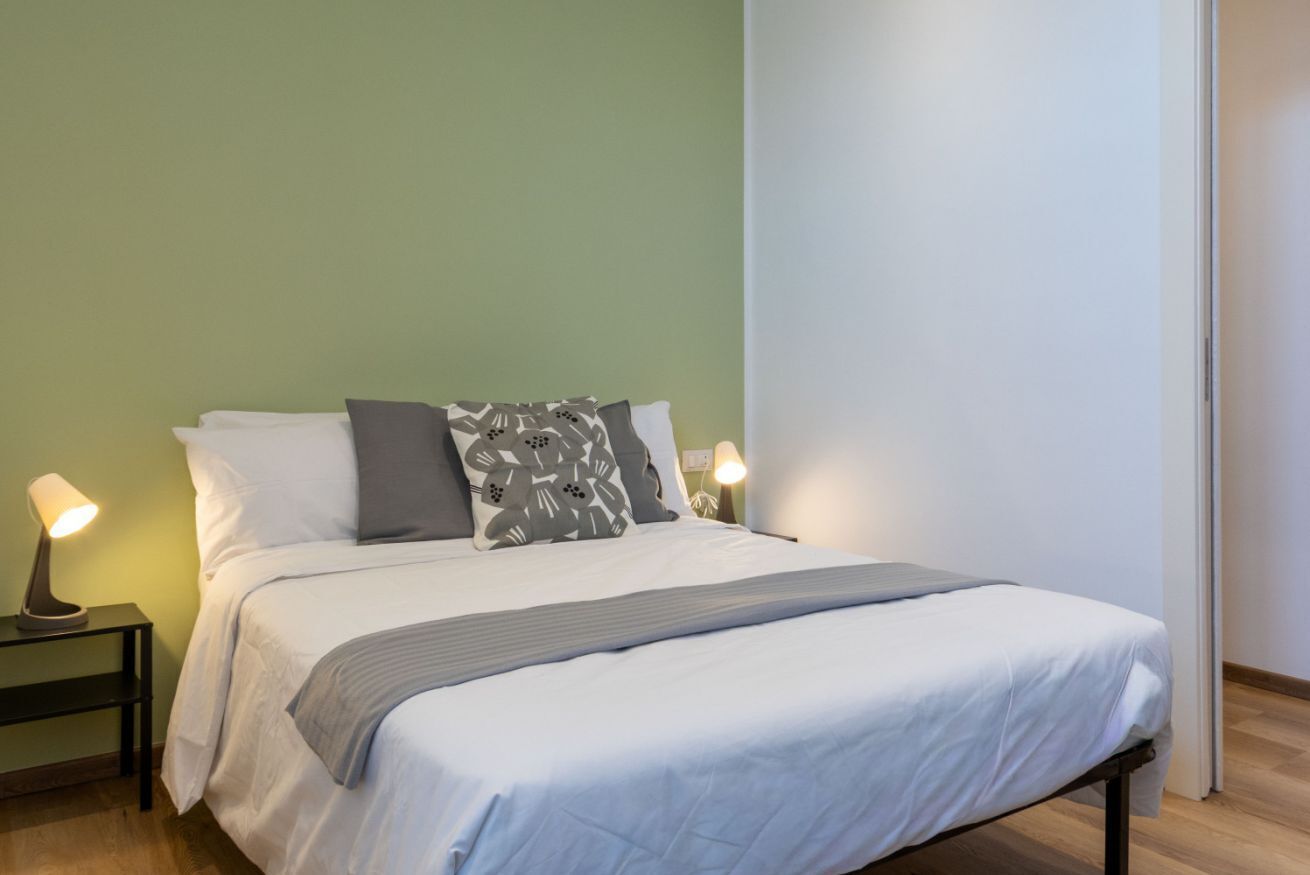 A neatly made bed with a white duvet and gray throw, two bedside lamps, and a green wall.