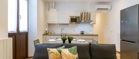 Una cocina moderna con armarios blancos, un refrigerador de acero inoxidable y un rincón comedor con sofá y cojines.