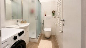 Ein modernes Badezimmer mit einer gläsernen Duschkabine, einem weißen Waschbecken, einer Toilette und einer Samsung Waschmaschine.