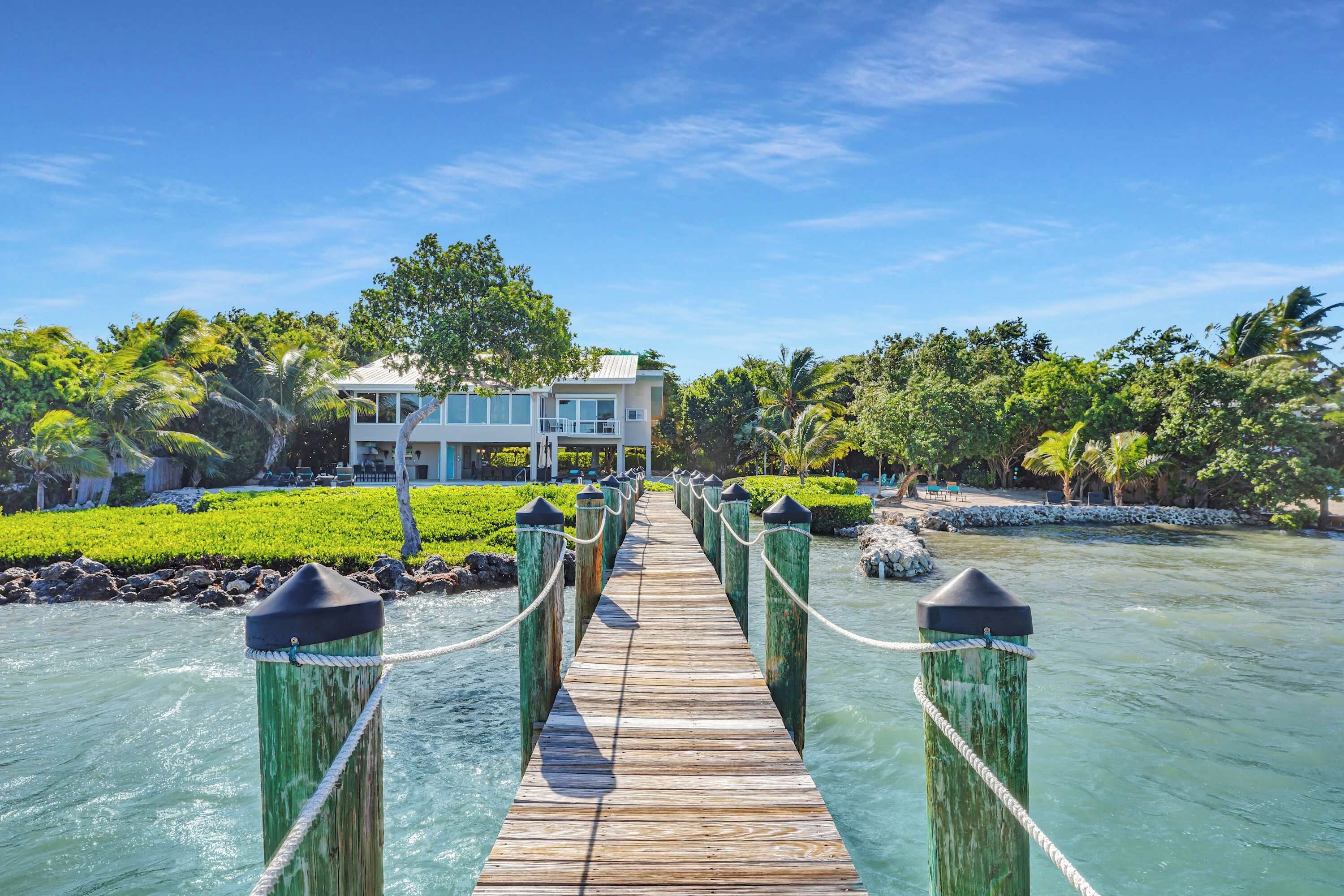 A wooden bridge leading to a modern building with a pool, surrounded by lush greenery and palm trees.