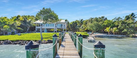 A wooden bridge leading to a modern building with a pool, surrounded by lush greenery and palm trees.