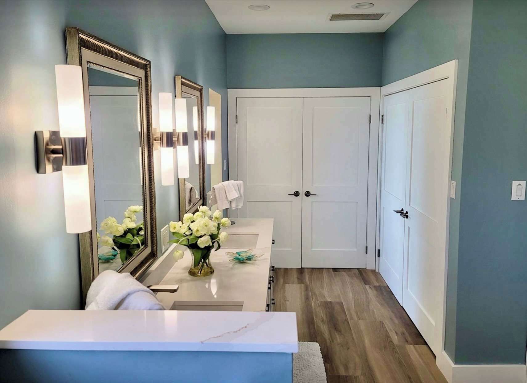 A bathroom with a vanity, two mirrors, a white door, and a vase with flowers.