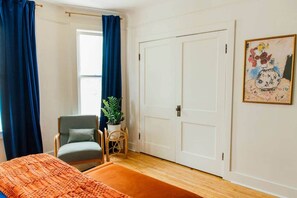 Ein Schlafzimmer mit Bett, einem Sessel, einer Zimmerpflanze, einer Tür und einem Bild an der Wand.