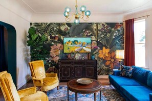 A living room with a large flat-screen TV, a blue sofa, a yellow armchair, a wooden cabinet, and a patterned rug. - Frida's House, Art-Filled Victorian in the Heart of Detroit (Detroit)