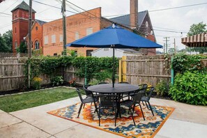 Eine Außenpatio mit rundem Tisch und Stühlen, einem blauen Sonnenschirm und einem gemusterten Teppich.