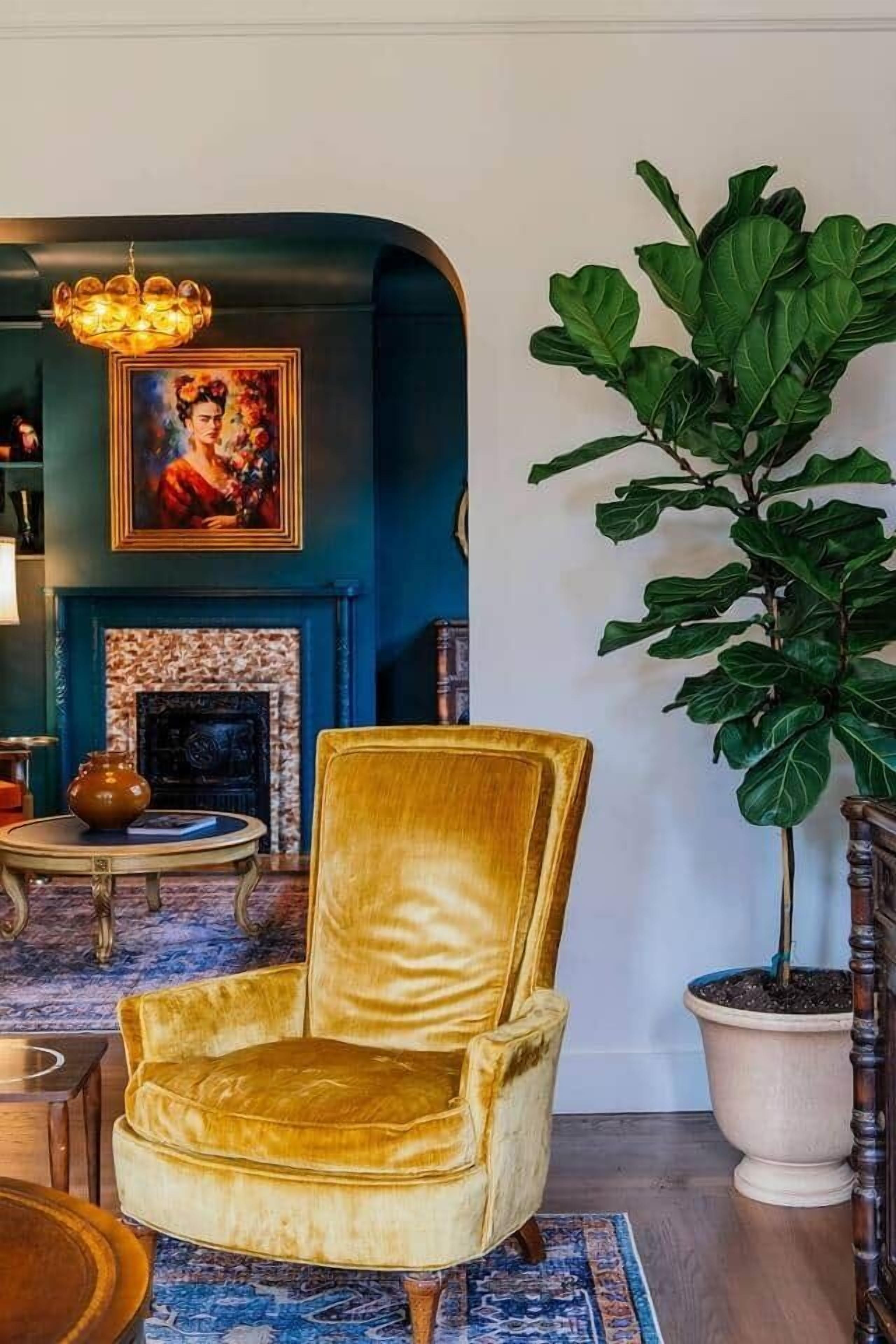 Een kamer met een grote potplant, een gouden fauteuil, een open haard en een schilderij aan de muur.