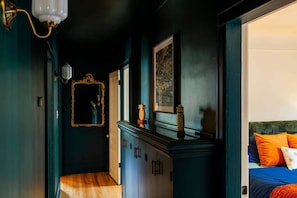 A hallway with a wooden floor, a dark wall, a mirror, and a bed with orange pillows. - Frida's House, Art-Filled Victorian in the Heart of Detroit (Detroit)