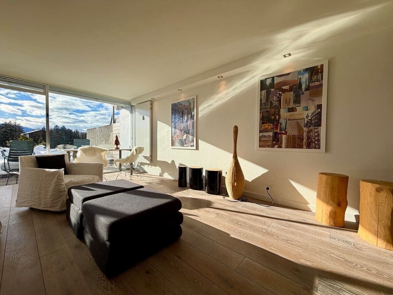 A modern living room with a sofa, a coffee table, and a wooden floor. There are two paintings on the wall and a window with a view of the sky.