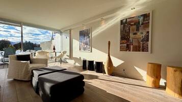 A modern living room with a sofa, a coffee table, and a wooden floor. There are two paintings on the wall and a window with a view of the sky.