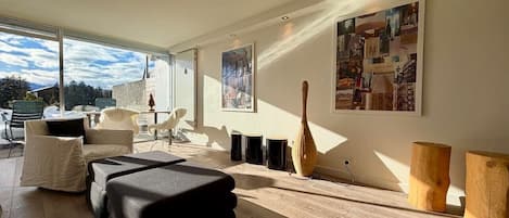 A modern living room with a sofa, a coffee table, and a wooden floor. There are two paintings on the wall and a window with a view of the sky.