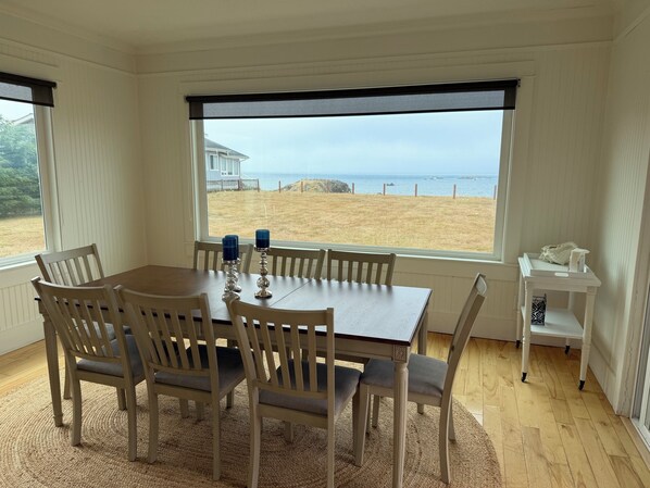 Un coin repas avec une table et des chaises, une vue sur la plage et une petite table d’appoint blanche sur laquelle se trouve une boîte de mouchoirs.