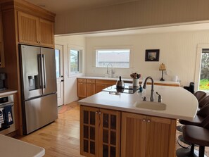 A modern kitchen with wooden cabinets, a stainless steel refrigerator, a built-in sink, and a dining area with a chair. - The Lighthouse! Ocean front bluff. (Crescent City)